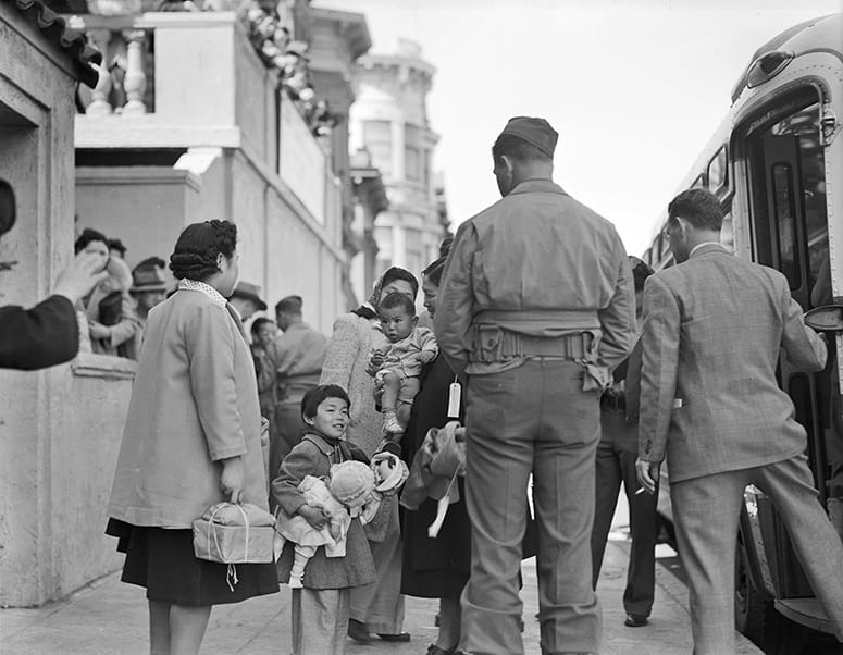Historic photo of a Japanese American mother and two children, next to two soldiers, about to board a bus.