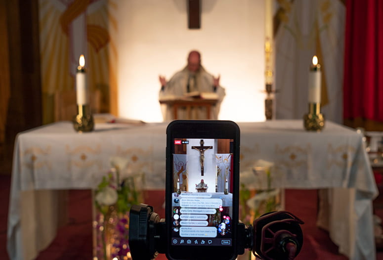 Photo of a phone set up to livestream a priest behind an altar, with text comments popin g up on the phone screen