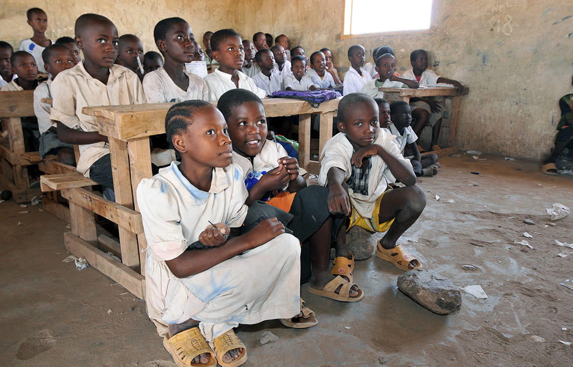 Children in a refugee camp schoolroom in Africa, some at desks some on the dirt floor
