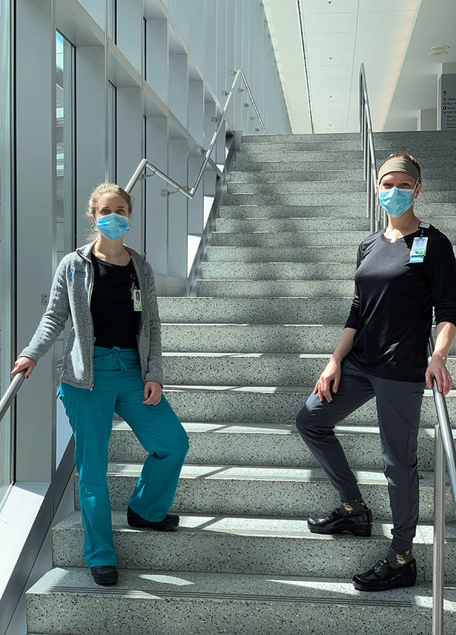 Two women dressed in scrubs, on the steps inside a hospital