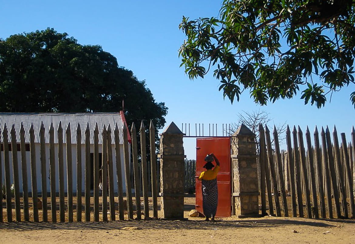 Photo of an African woman at the gate of a village surrounded by a loose wooden fence