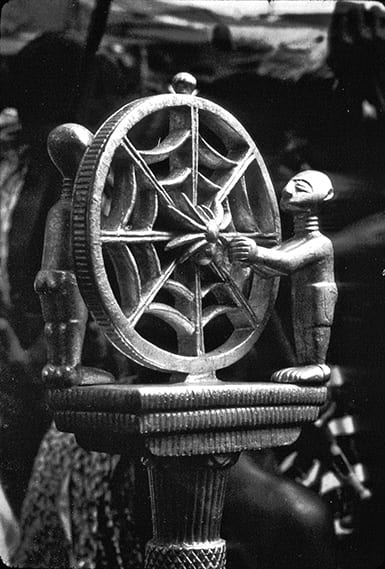The top of a wooden divination staff carved as two figures next to a spider web with a spider in the center