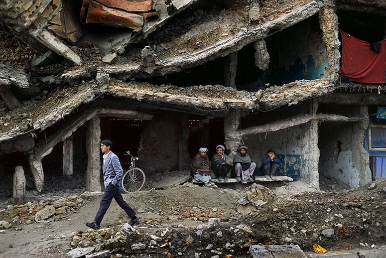 Photo of four Afghani men sitting in the ruined shell of a building while another man walks past, on the rubble-covered street