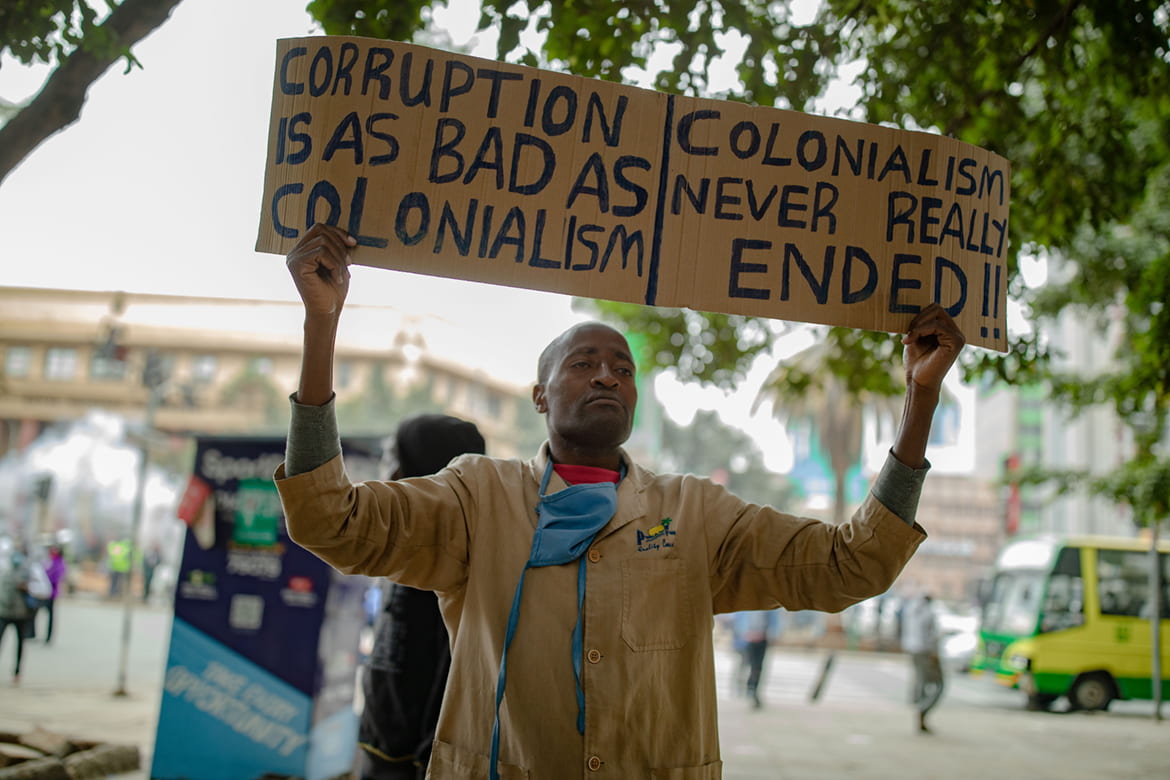 Man holding a protest sign reading Corruptions is as bad as colonialism. Colonialism never really ended.