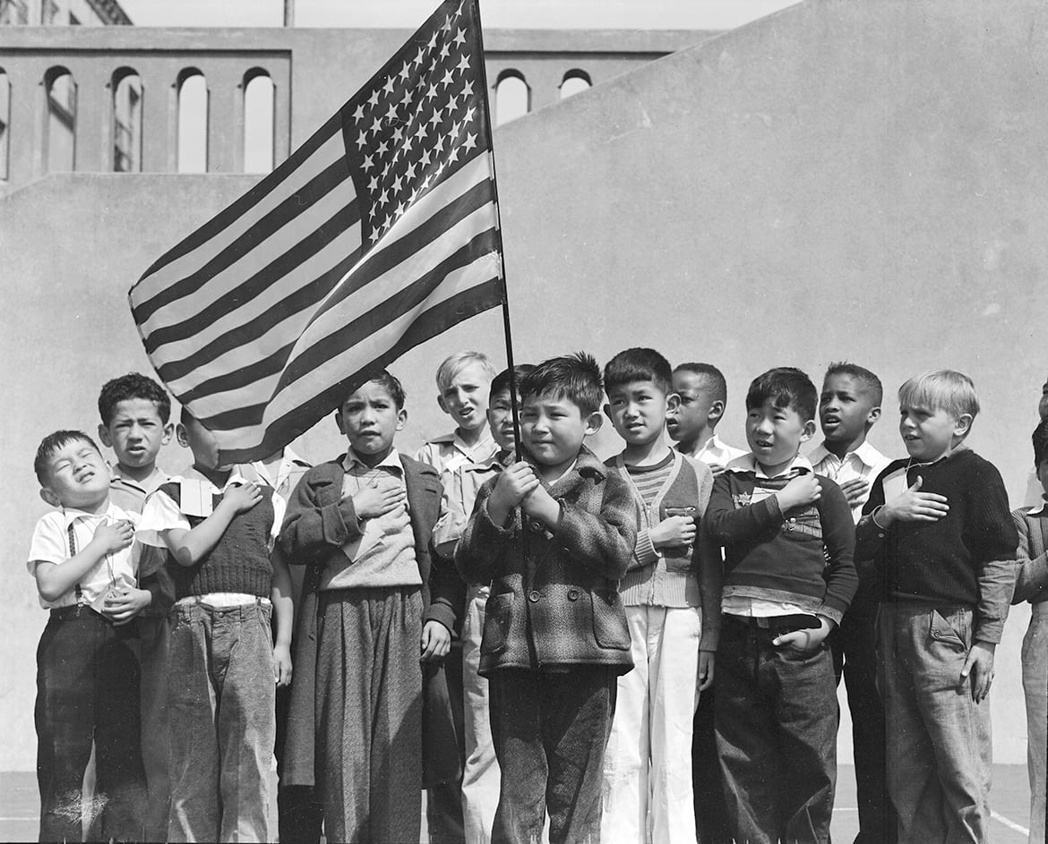 1942 photo of young students holding an American flag and holding hands over their hearts.