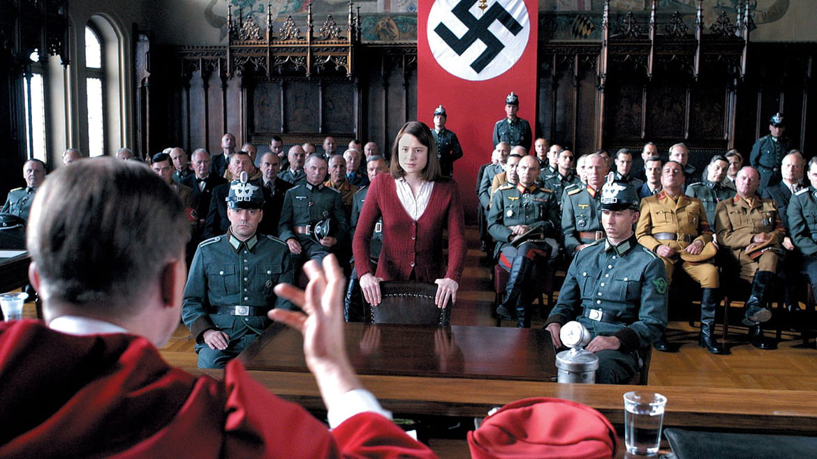 Woman standing in a courtroom full of soldiers with a Nazi flag hanging in the back