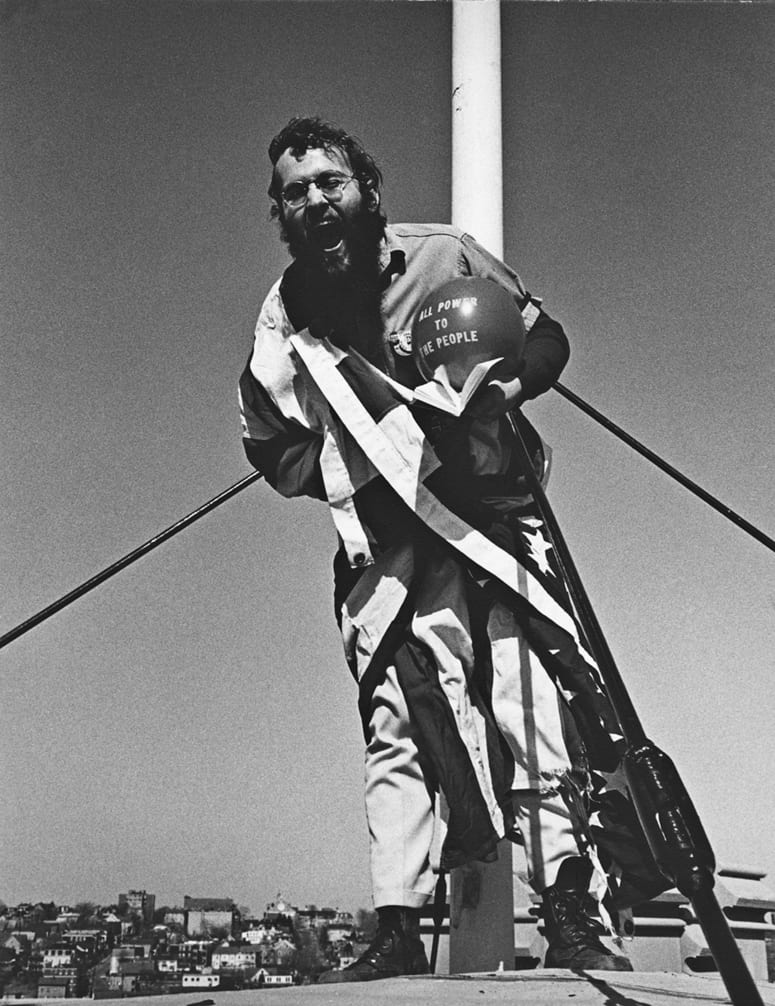 Man draped in American Flag, leaning away from flag pole, yelling towards the camera
