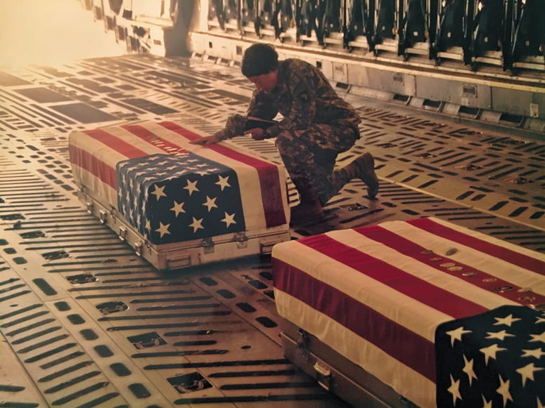 Military chaplain holding a bible and kneeling next to flag-draped coffins in the back of an airplane