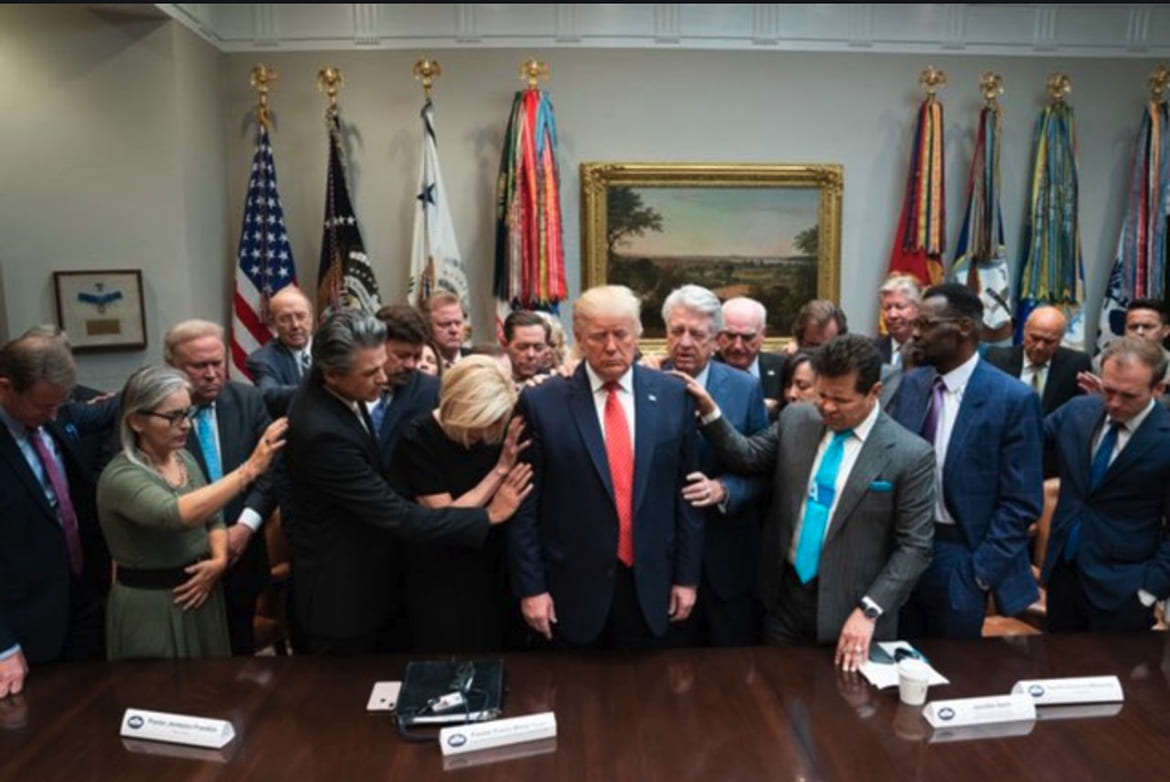 Ministers surrounding Trump with their hands placed on each others shoulders and heads bowed in prayer