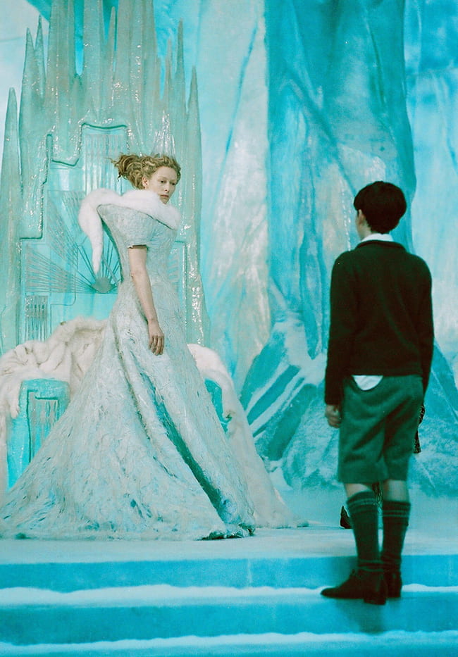 Woman in an ornate gown looking at a boy standing at attention, both inside an ice cavern