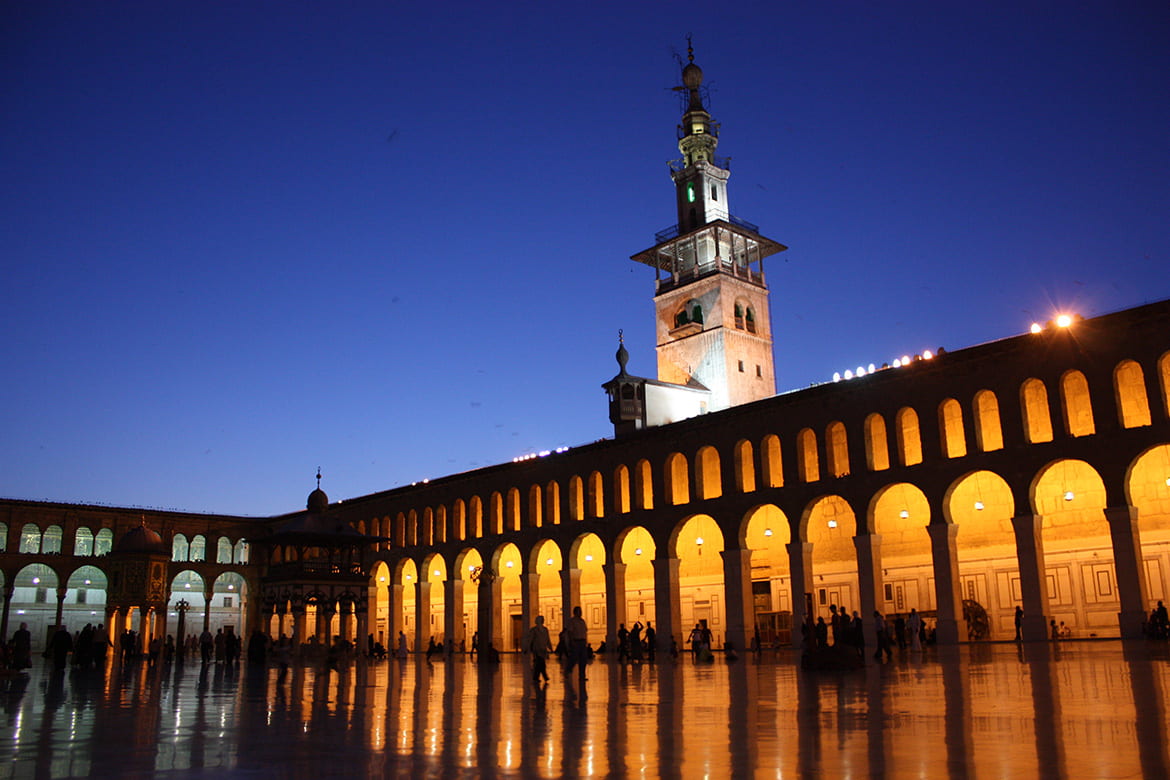 A courtyard inside the Umayyad Mosque, with walls filled with arches, lit up at night 