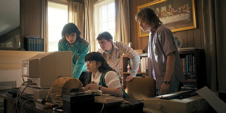 Four teenagers clusters around a 1980s computer, a painting of the Last Supper on the wall behind them
