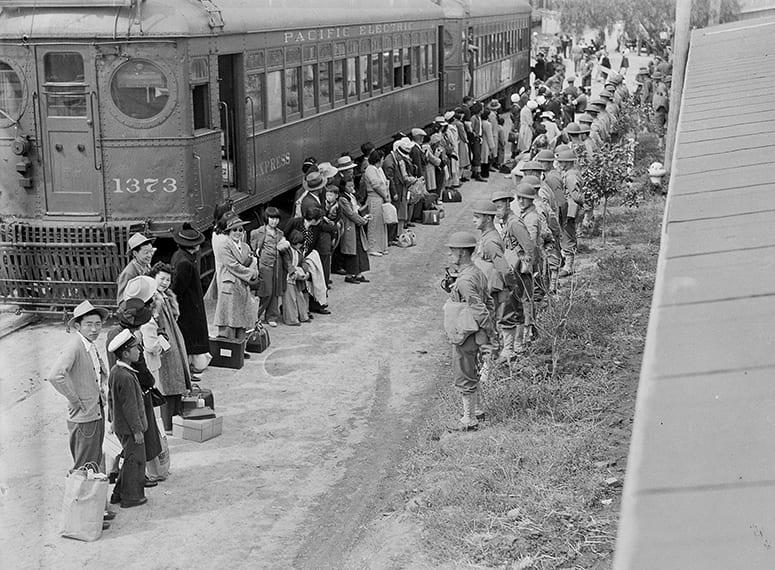 Historic photo of Japanese Americans lined up next to a train, facing a line of soldiers.