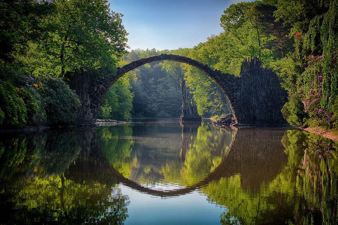 Photo of a lake in a green forest, with a stone arch bridge reflected in the still water