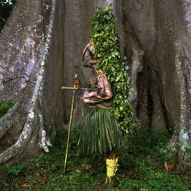 Person wearing a costume of leaves and cloth, standing in front of a massive tree trunk.