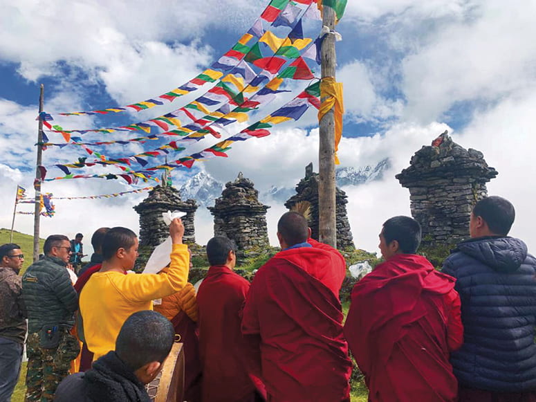 Buddhist monks and other participants gathered outside by stone stupas, under prayer flags