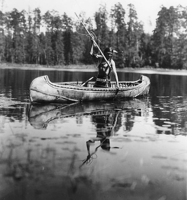 Historic photo of a Native American fisherman in a canoe, holding his spear over the water